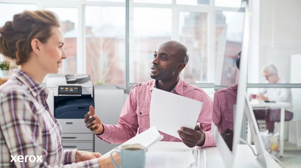 Xerox® Workplace Cloud und Suite: Zentralisiertes Flottenmanagement Eine Person überprüft Daten auf einem Laptop-Bildschirm mit einer digitalen Überlagerung verschiedener Gerätesymbole (Drucker, Mobilgeräte) und verbundener Linien, die das zentralisierte Flottenmanagement für Xerox Workplace Solutions symbolisieren.