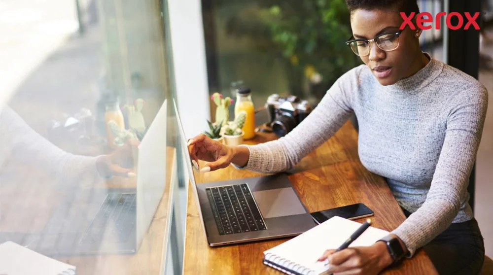 Eine Person mit Brille arbeitet an einem Holztisch, schaut auf einen Laptop-Bildschirm und schreibt in ein Notizbuch, während ein Smartphone in der Nähe ist, was auf eine produktive Arbeitsumgebung hindeutet