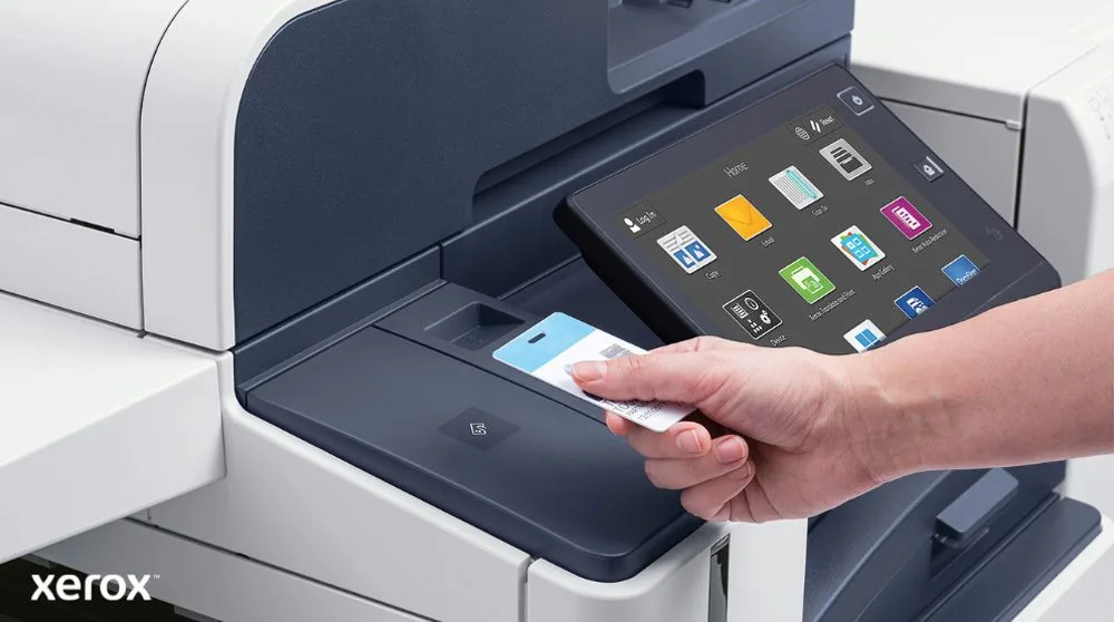 Xerox® Workplace Cloud und Suite: Sichere Authentifizierung und intelligente Schnittstelle Eine Hand hält eine Sicherheitskarte und scannt sie in der Nähe des Kartenlesers eines modernen Xerox-Multifunktionsdruckers mit einem großen Touchscreen-Display, auf dem verschiedene Anwendungssymbole angezeigt werden.