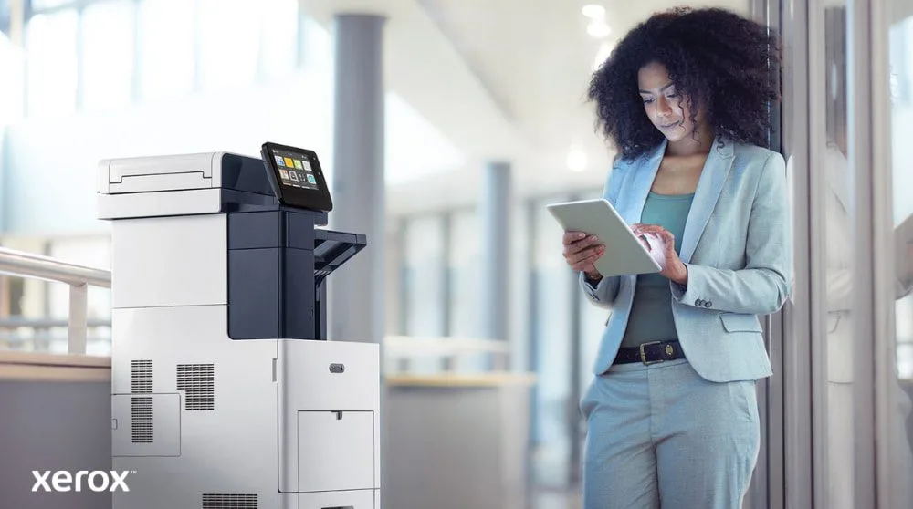 Eine Person steht neben einem Xerox VersaLink B605-Multifunktionsdrucker, betrachtet ein Tablet-Gerät und interagiert damit. Dabei wird veranschaulicht, wie ConnectKey die mobile Interaktion mit dem Drucker ermöglicht.